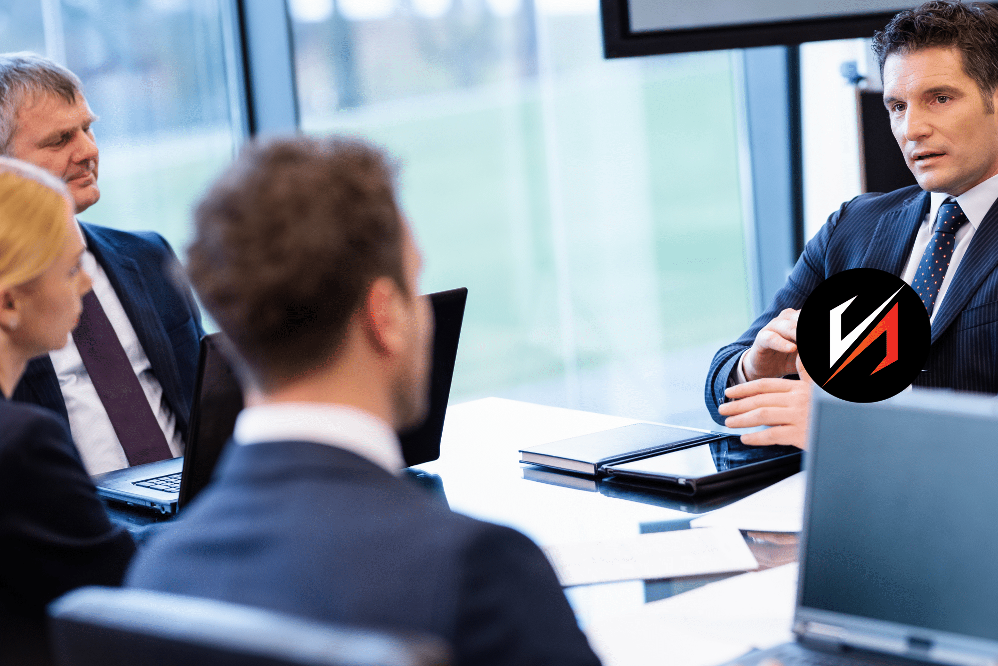 Business meeting with four professionals discussing digital marketing strategies, featuring a man in a suit gesturing, laptops and documents on the table, emphasizing leadership and marketing governance.